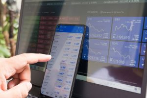 Person clicking on a financial app on their smartphone, with a similar app showing on a monitor behind the phone