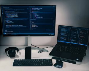 A PC monitor, laptop, keyboard placed on a desk, the display showing code