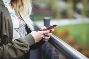 Gril with blonde hair in an olive jacket holds a red smartphone and looks down at the screen beside a railing, outdoors.